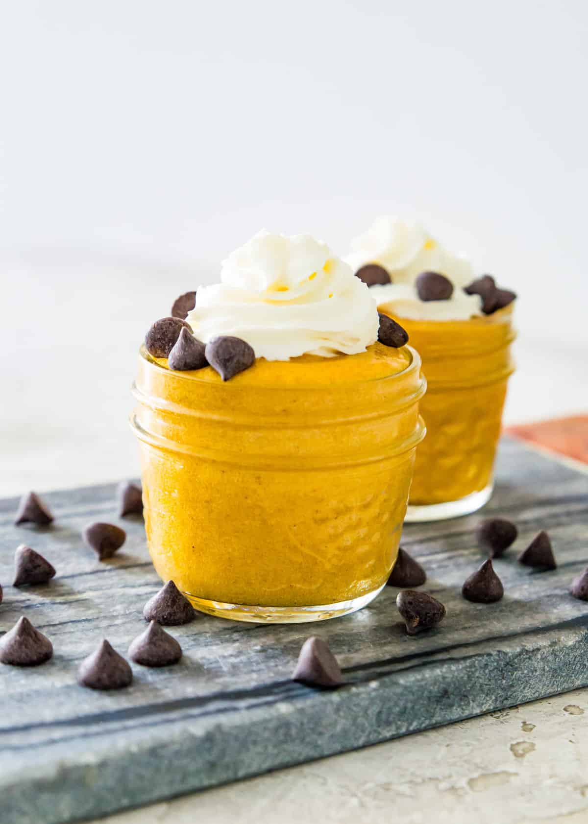 Two clear jars filled with pumpkin mousse topped with whipped cream and chocolate chips. 