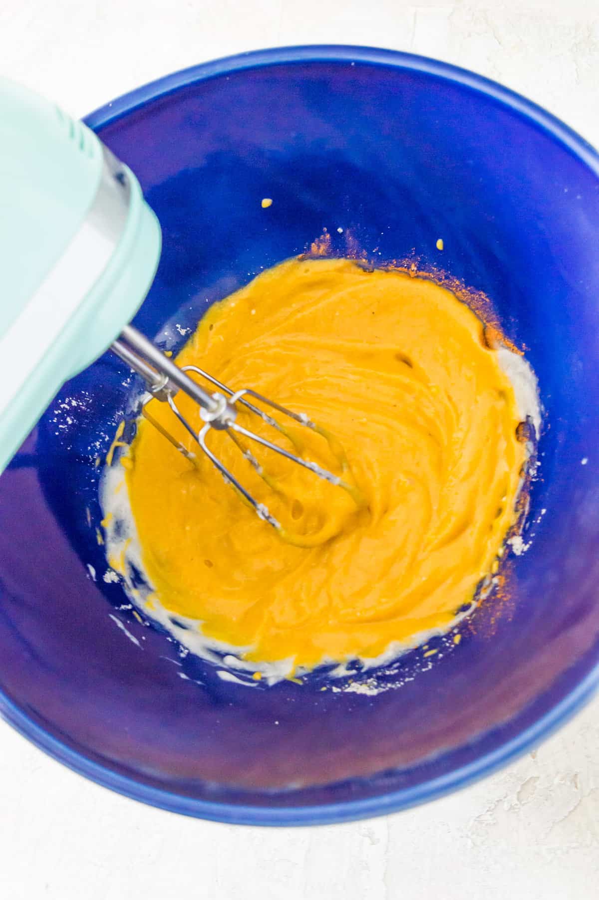 Pumpkin puree and cream cheese in a large blue bowl being blended together with a hand mixer. 