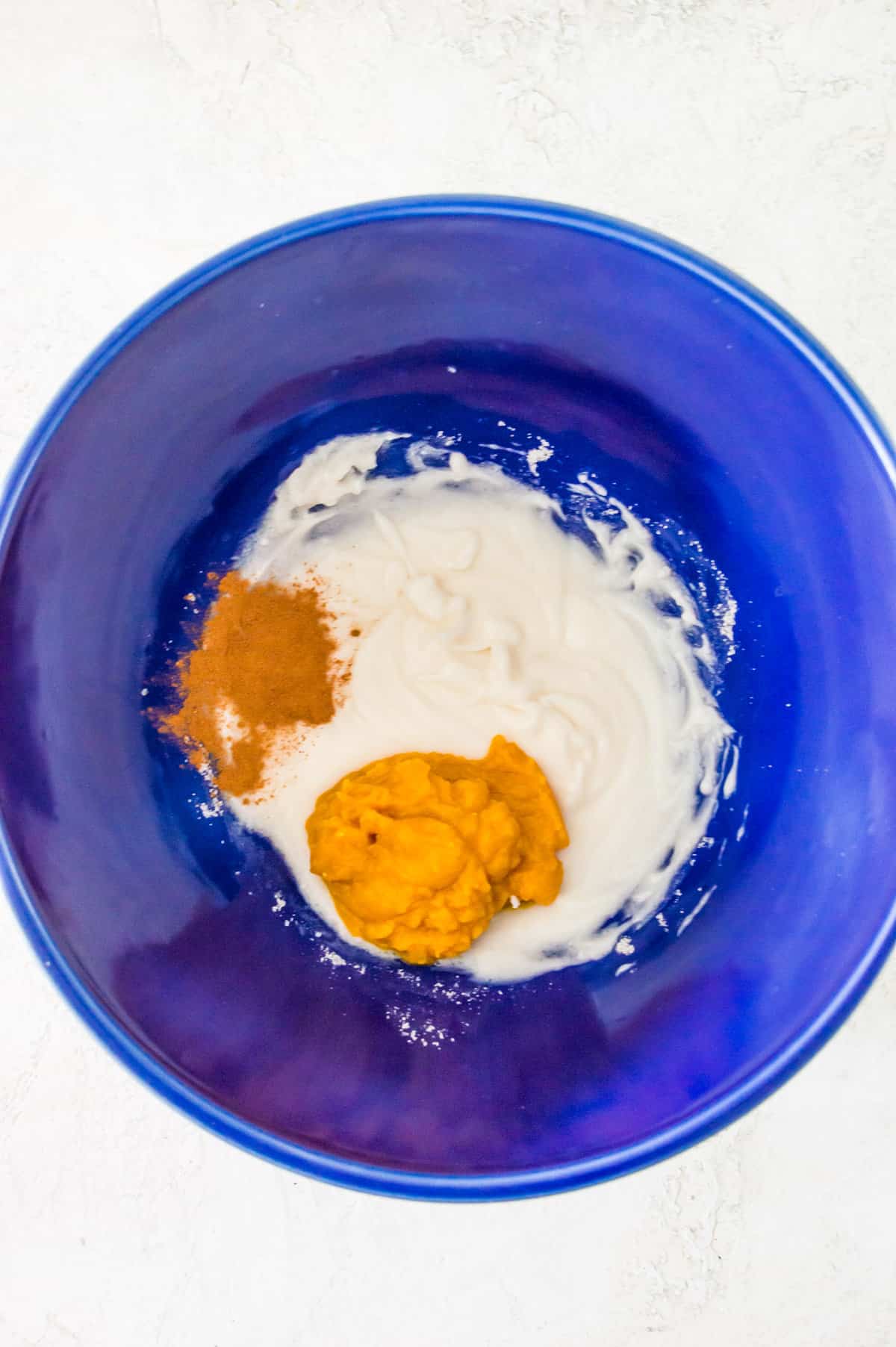 Blended cream cheese in a large blue bowl with a spoonful of pumpkin puree and pumpkin pie spice on top of it.