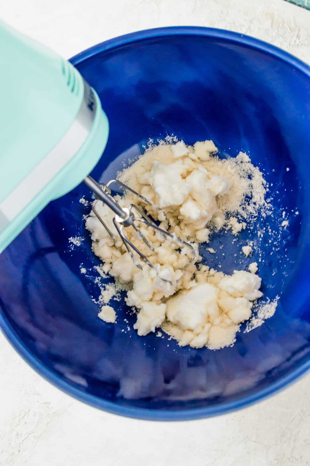 Cream cheese and cane sugar in a large blue bowl being blended with a hand mixer.