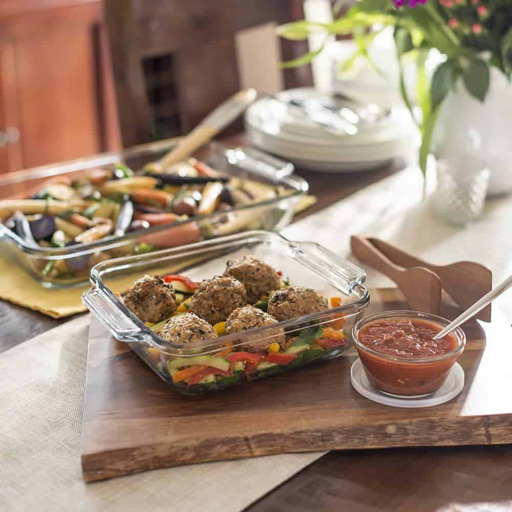 A few different glass casserole dishes filled with baked meatballs, roasted vegetables with wooden utensils next to them. 