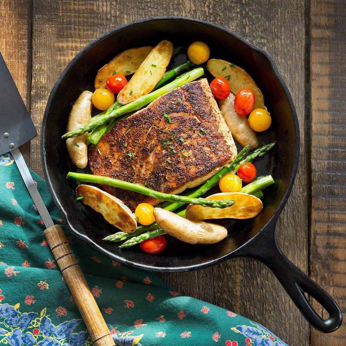 A Simple Chef cast iron skillet with roasted fish, potatoes, tomatoes and asparagus in it.