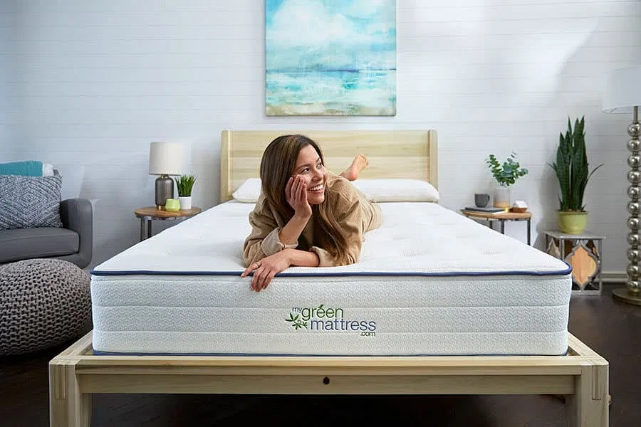 A woman with long brown hair laying on a mattress on a wooden bed frame. The bed has two side tables beside it and plants beside it.