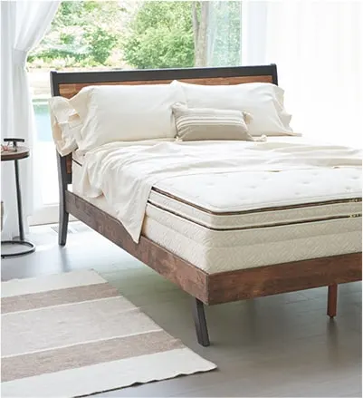 A wooden bed frame in front of a window with a Naturpedic brand mattress on it plus three pillows. 