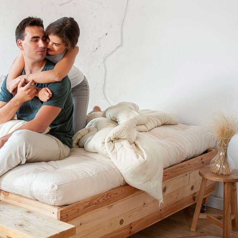 A man and woman hugging on a white mattress with a wooden beside table beside them.