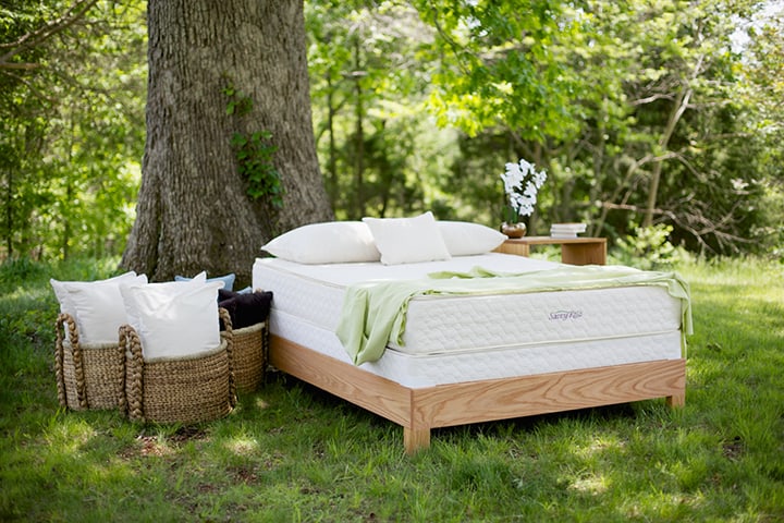 A wooden bed frame with a mattress on it in the forest with a large tree beside it plus baskets of pillows around it.