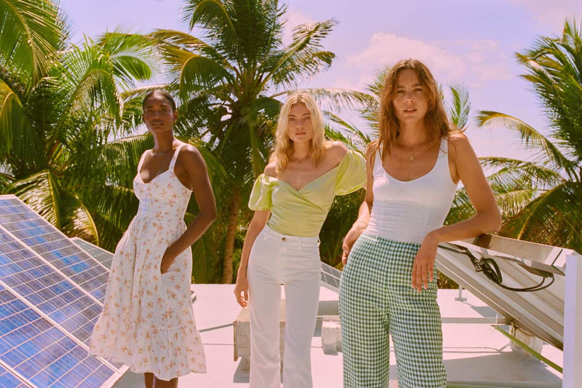Three girls on a rooftop surrounded by palm trees with one girl wearing a sundress, one in a tank top with checked pants and the other in a v-neck top with white jeans.