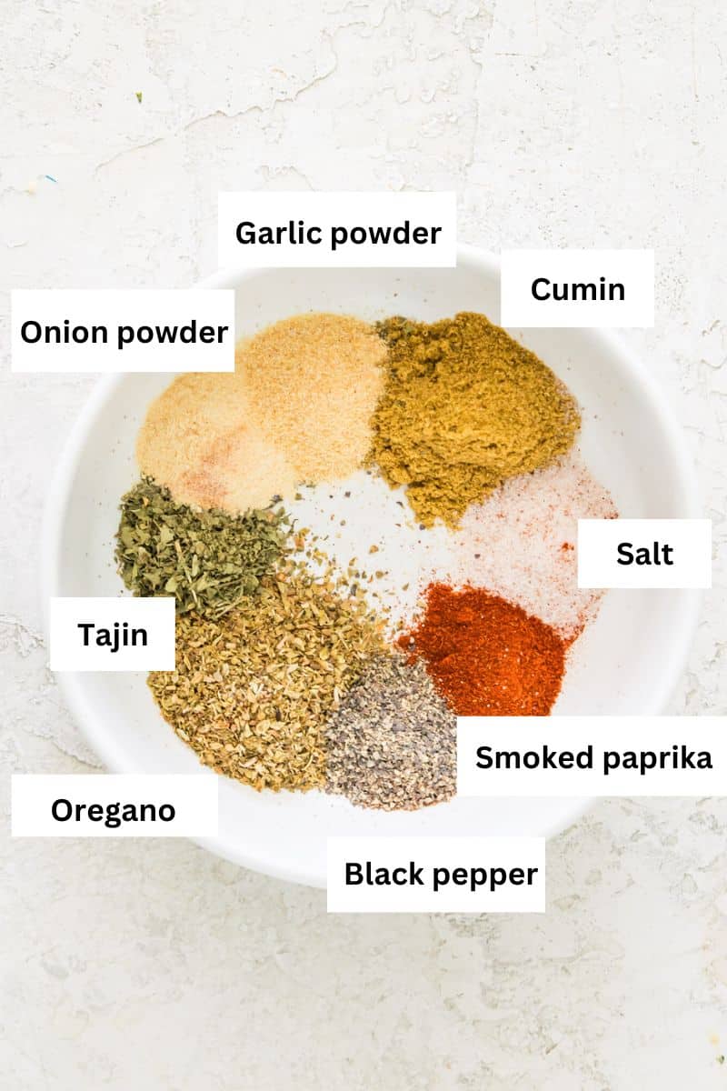A white bowl filled with different dried herbs and spices, including black pepper, oregano, paprika, salt, onion powder, garlic powder and cumin.