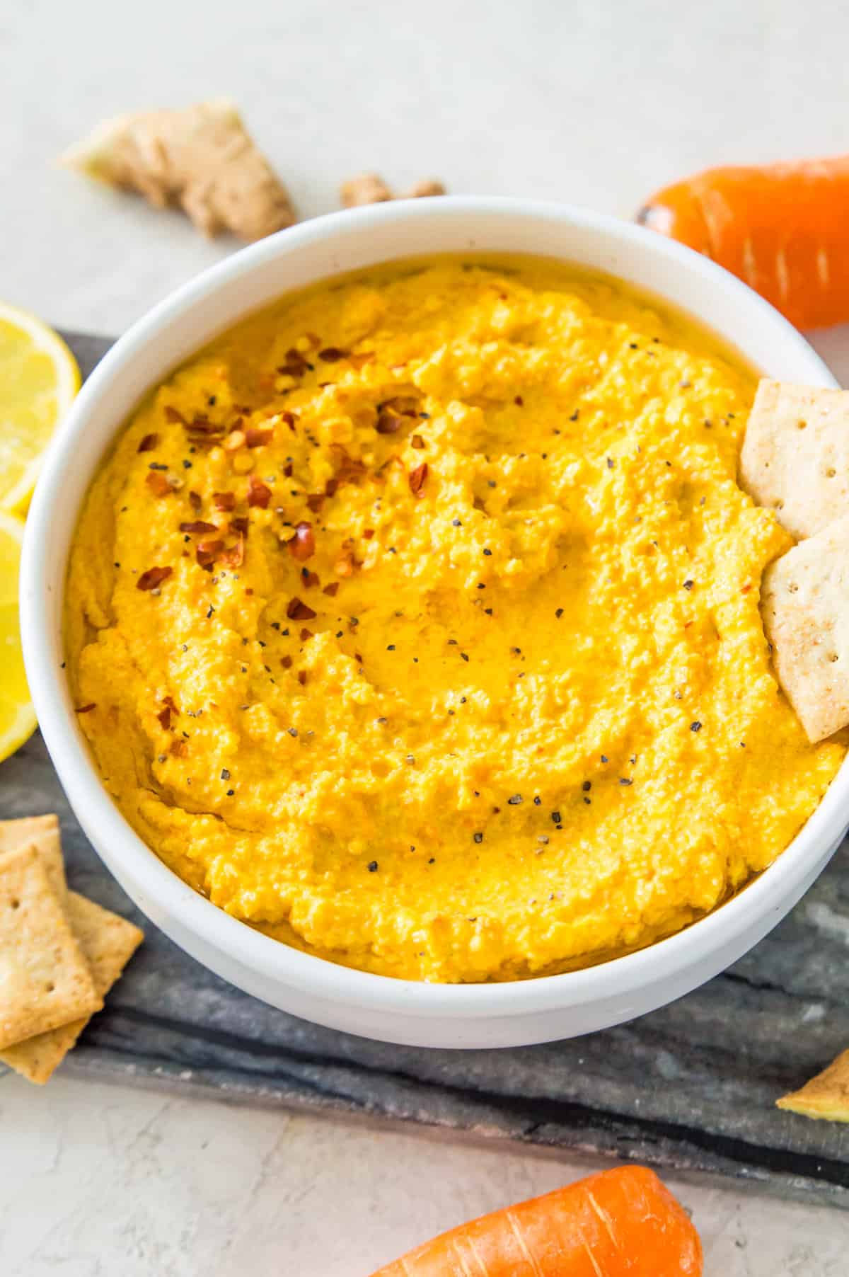 A bowl filled with a carrot dip topped with chili flakes with two crackers in it.