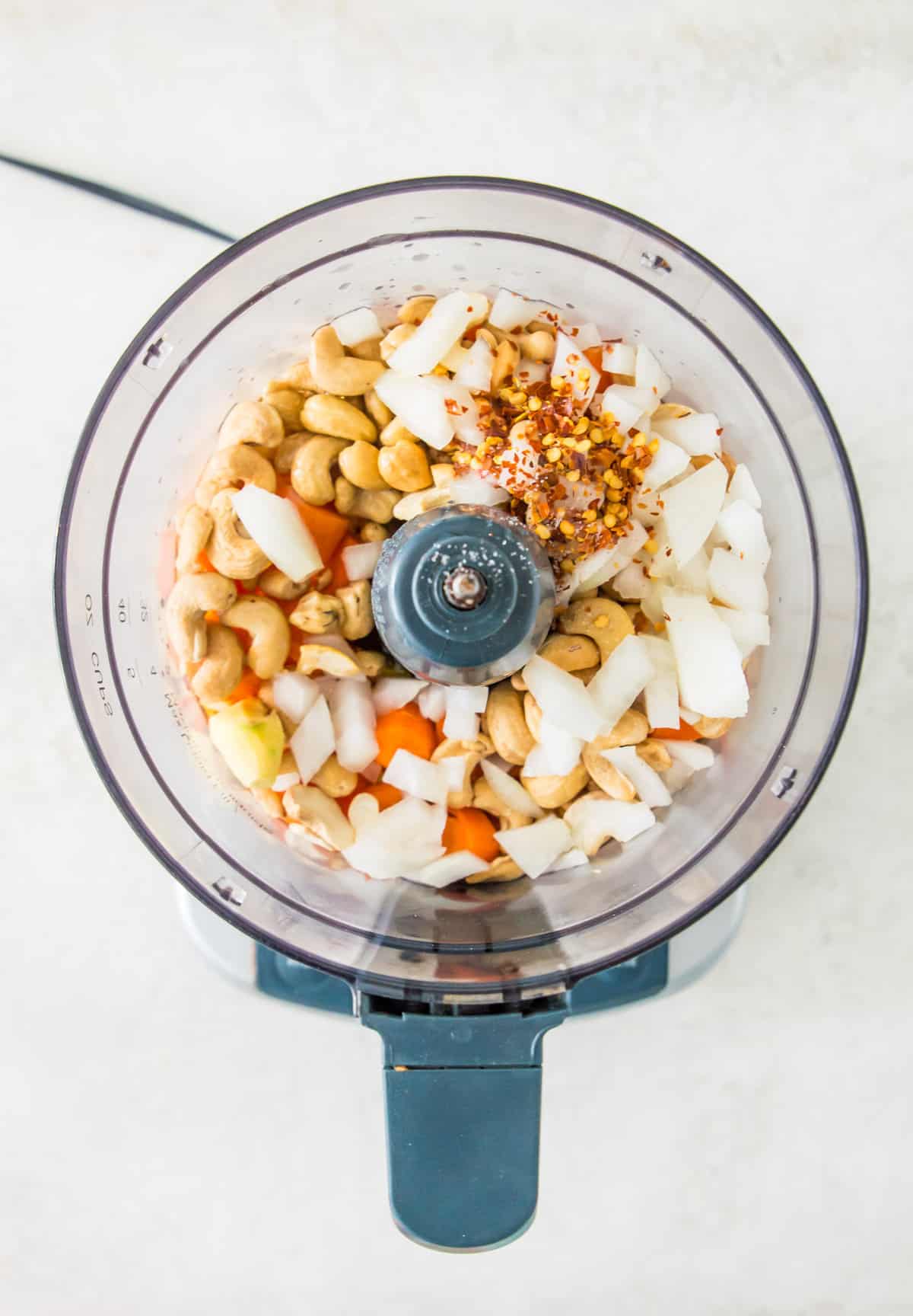 A food processor filled with cashews, chopped onion, chopped carrots and spices. 