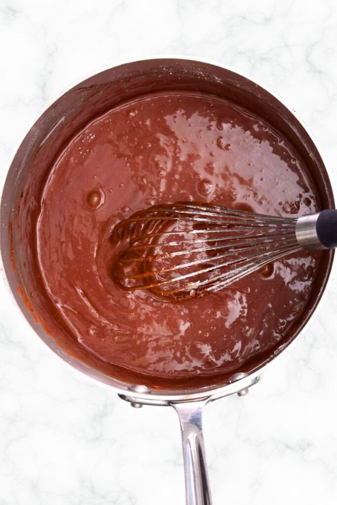 A pot with chocolate icing in it with a whisk stirring the icing. 