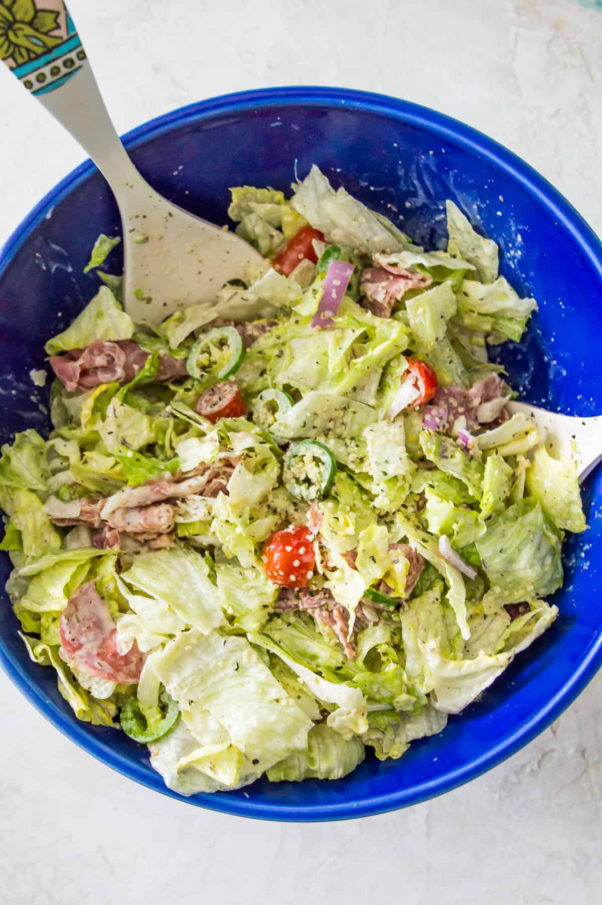 A large blue bowl with a grinder salad in it made with shredded lettuce, grape tomatoes, salami and banana peppers. 