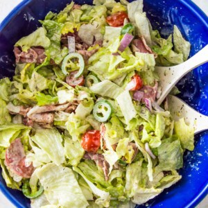 An Italian grinder salad in a large blue bowl topped with cherry tomatoes, chopped banana peppers and parmesan cheese.