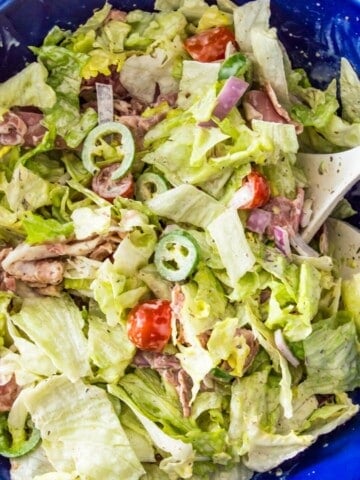An Italian grinder salad in a large blue bowl topped with cherry tomatoes, chopped banana peppers and parmesan cheese.