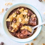 A snickers mug cake in a mug, topped with a caramel sauce, chocolate chips and peanuts with a spoon in it.