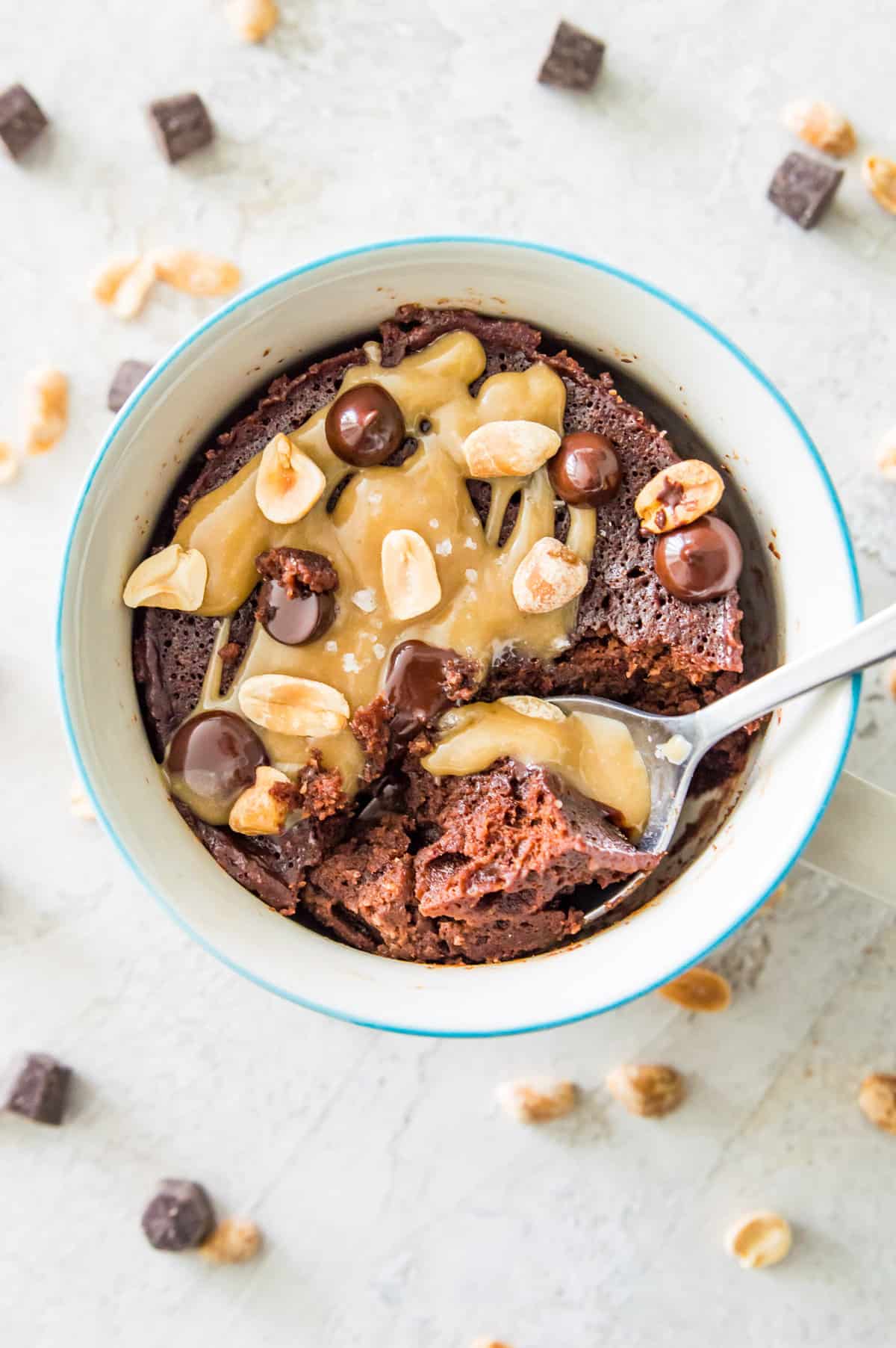 A chocolate mug cake topped with caramel sauce, peanuts and chocolate chips with a spoon taking a piece out of it.