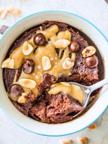 A snickers mug cake in a mug, topped with a caramel sauce, chocolate chips and peanuts with a spoon in it.