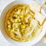 A bowl filled with a vegan dill pickle dip topped with chopped dill pickles and two crackers.