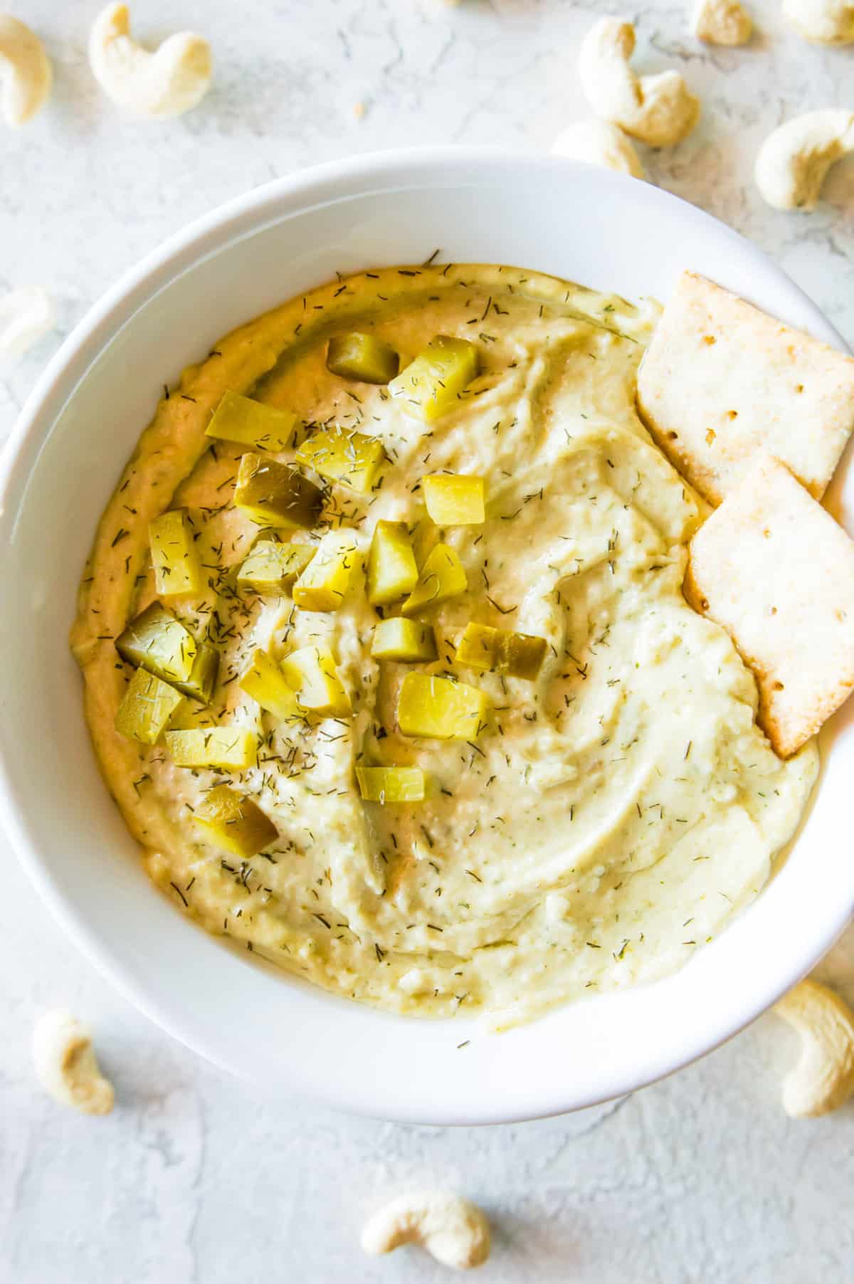 A bowl of vegan dill pickle dip topped with chopped dill pickles, fresh dill and with two crackers in it.