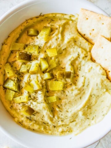 A bowl filled with a vegan dill pickle dip topped with chopped dill pickles and two crackers.
