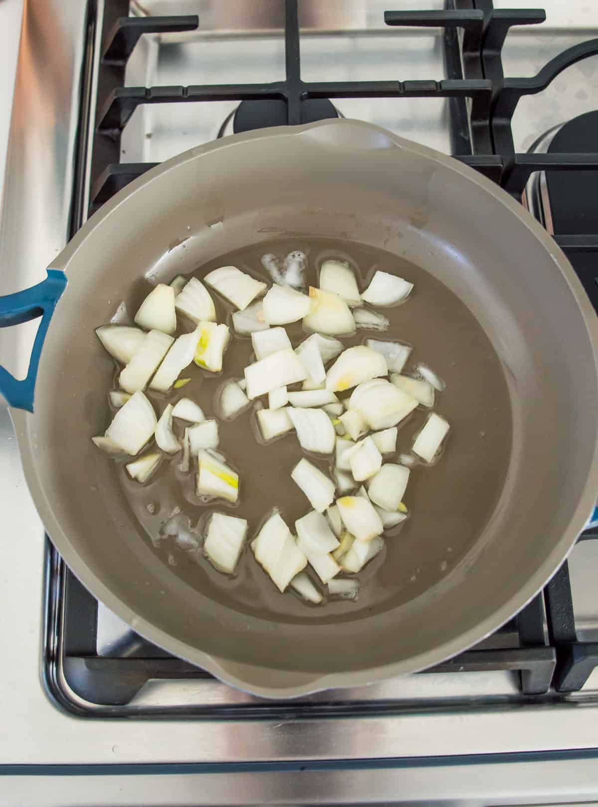 A grey pan on the stovetop with chopped onion cooking in it.
