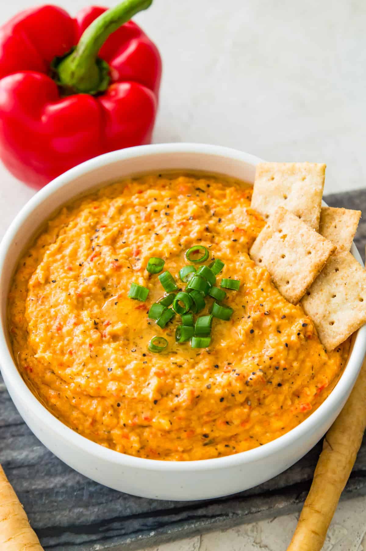 A bowl of vegan roasted red pepper dip garnished with chopped green onion with four crackers in it.