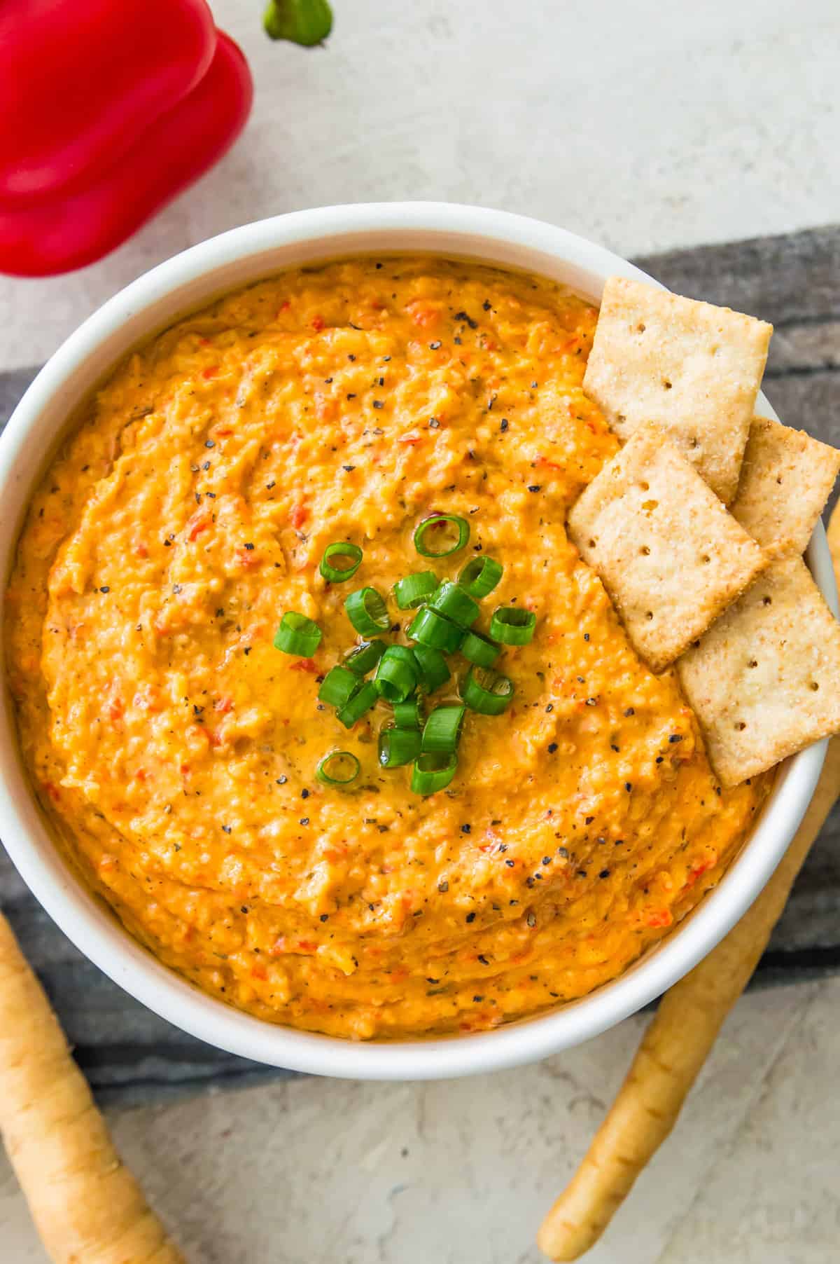 A bowl with a roasted red pepper dip in it topped with chopped green onion and with crackers in it.