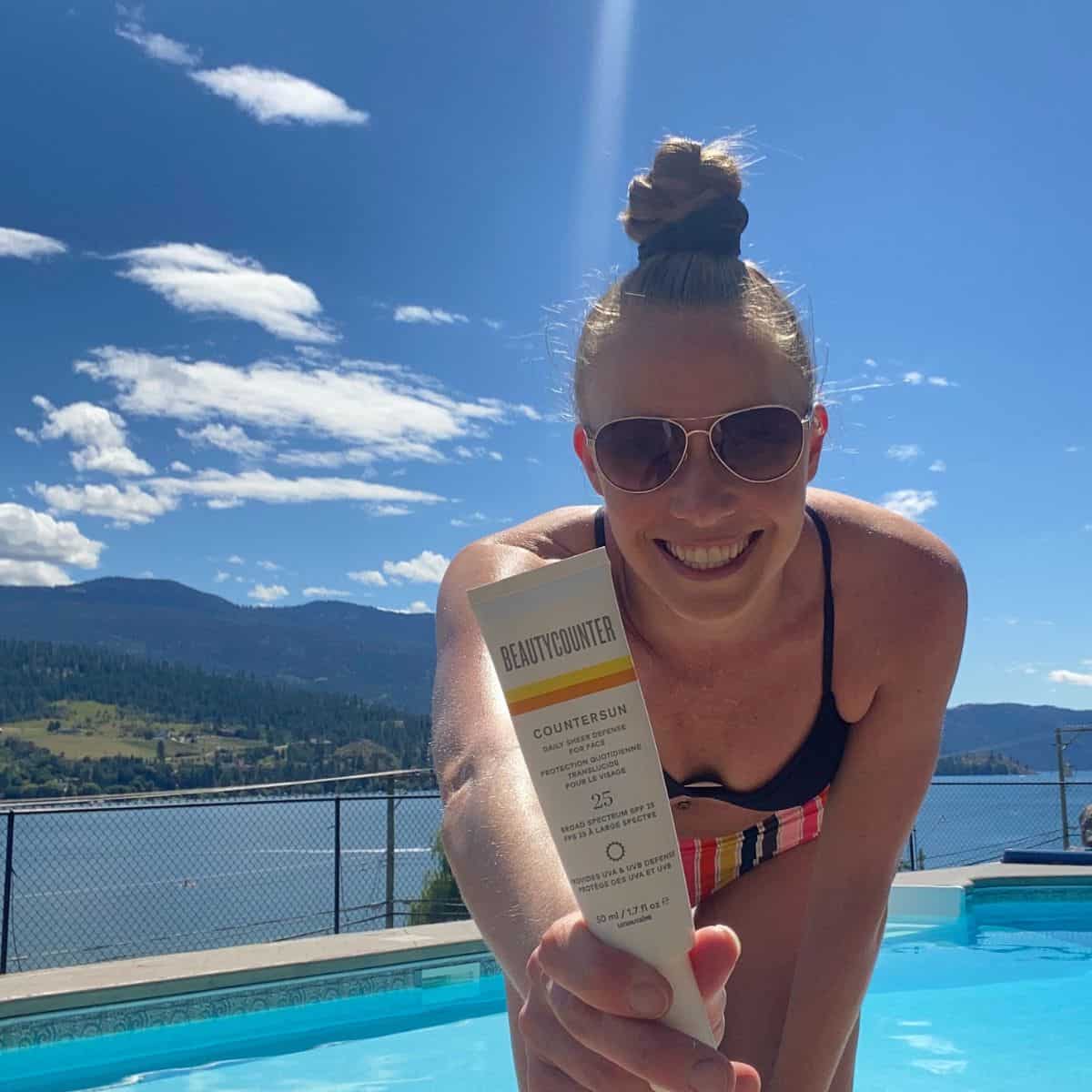 A girl in a bikini with a pool behind her holding a tube of Beautycounter sunscreen.