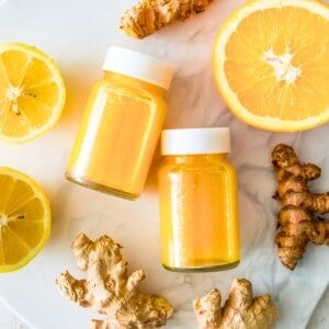 Two ginger turmeric shots in glass bottles with white lids surrounded by ginger, turmeric, orange slices and lemon slices.