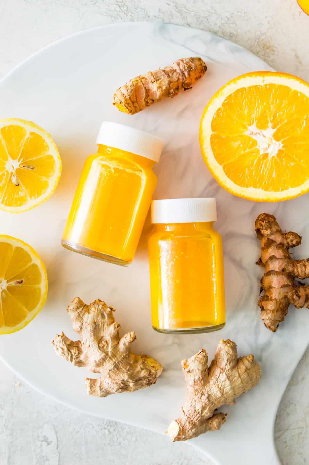 Two small glass bottles filled with a ginger turmeric drink surrounded by pieces of fresh ginger, turmeric and orange slices.