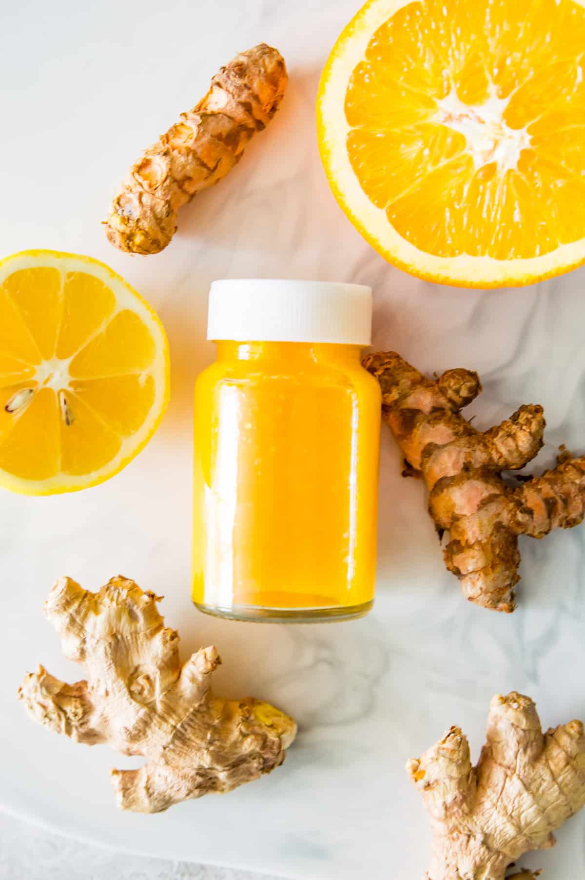 A small glass bottle filled with a ginger wellness drink surrounded by lemon slices, orange slices, fresh ginger and fresh turmeric.