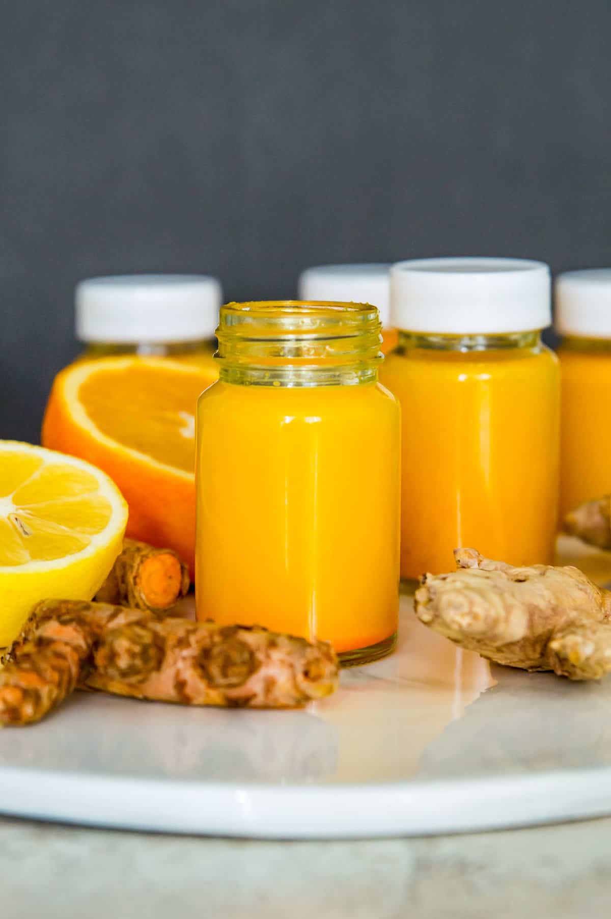 Small glass bottles filled with an orange liquid surrounded by fresh ginger, turmeric, orange slices and lemon slices.