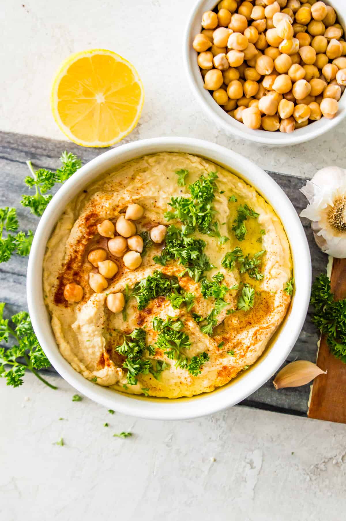 A bowl filled with a gluten free hummus that is topped with fresh, chopped parsley and chickpeas and the bowl has a lemon wedge and garlic cloves beside it.