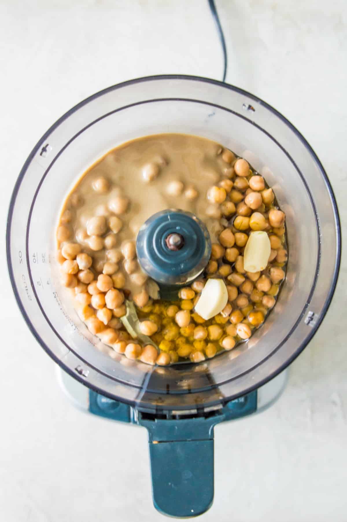 A food processor filled with ingredients for making hummus including chickpeas, oil, tahini, garlic cloves and spices.