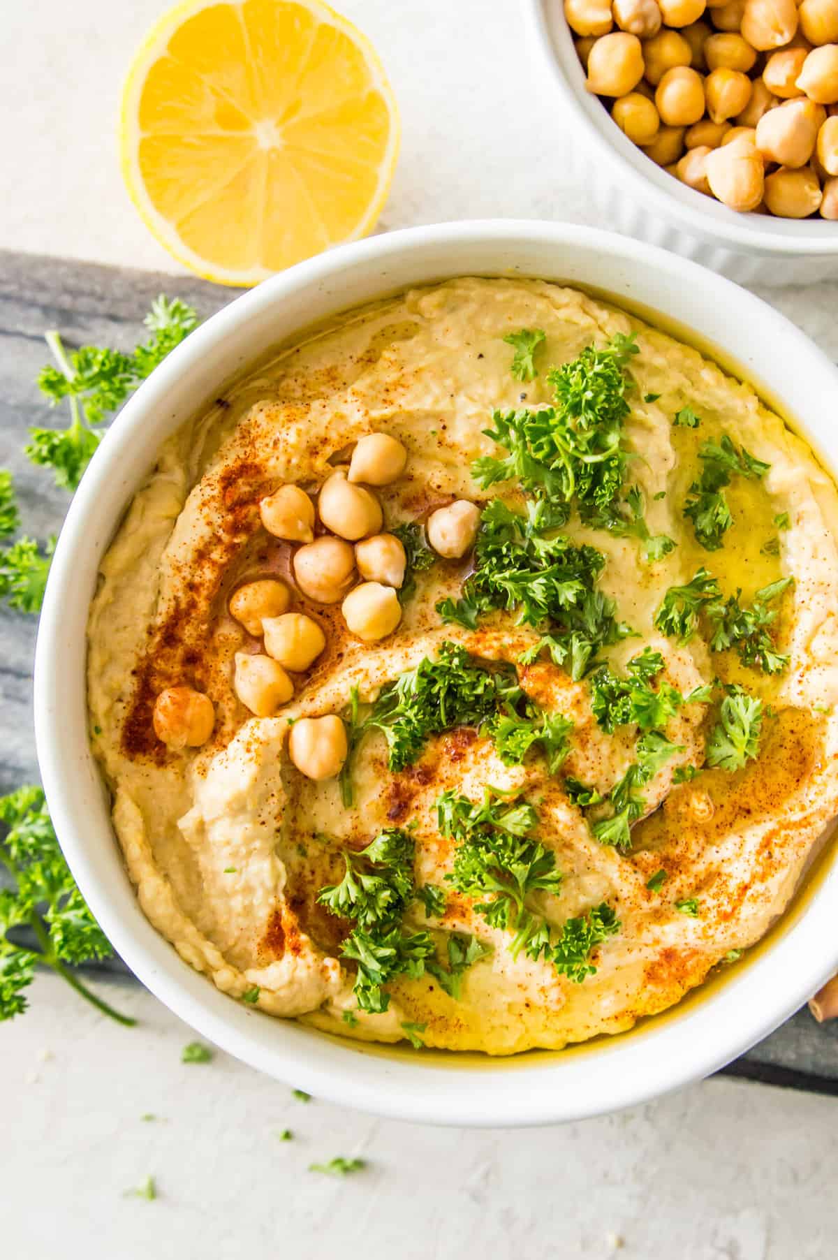 A bowl filled with hummus topped with chopped fresh parsley, paprika and chickpeas surrounded by a lemon wedge and a bowl of chickpeas.