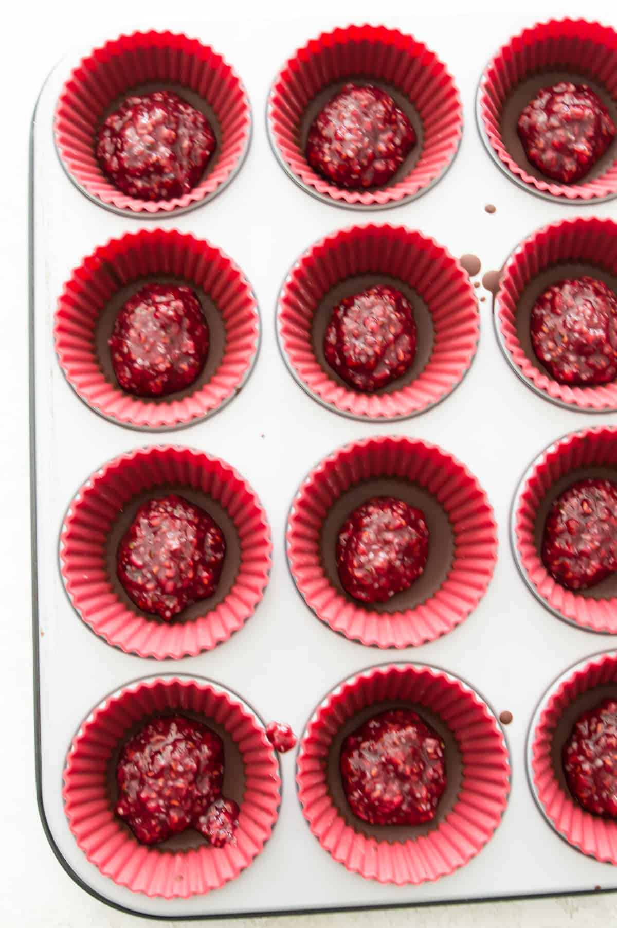 A muffin tray lined with silicone muffin cups and each one has some chocolate in it topped with a raspberry filling.