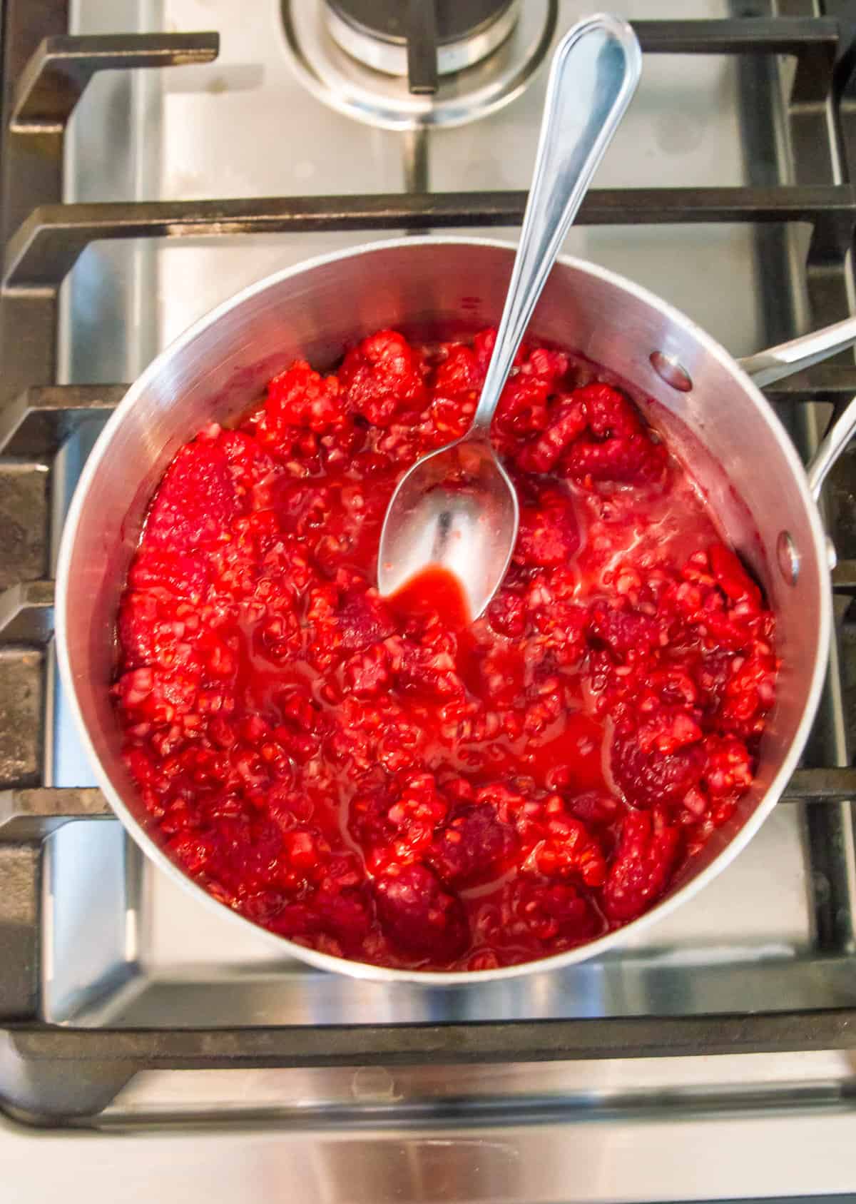 A pot on the stovetop with broken up raspberries in it with a spoon in it.