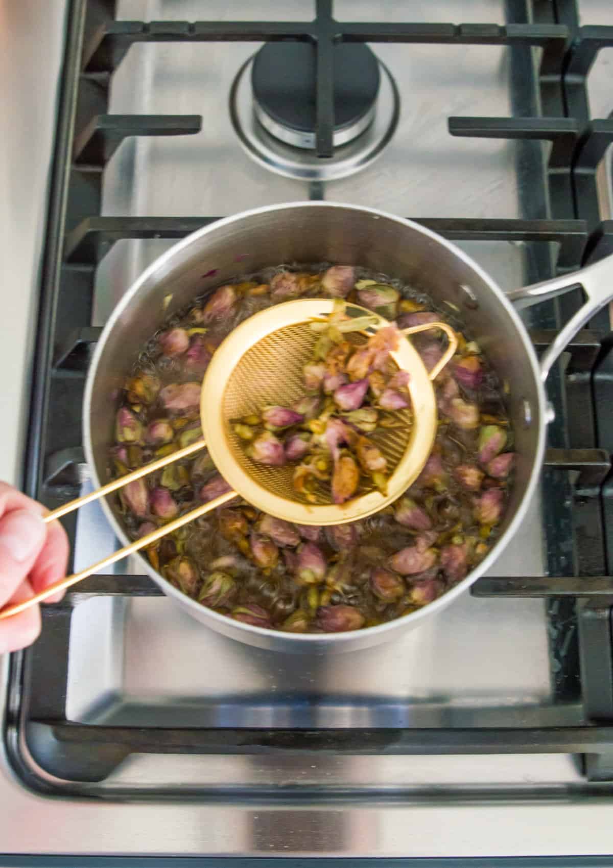 A pot filled with water and rosebuds with a small gold strainer scooping rosebuds out of it.