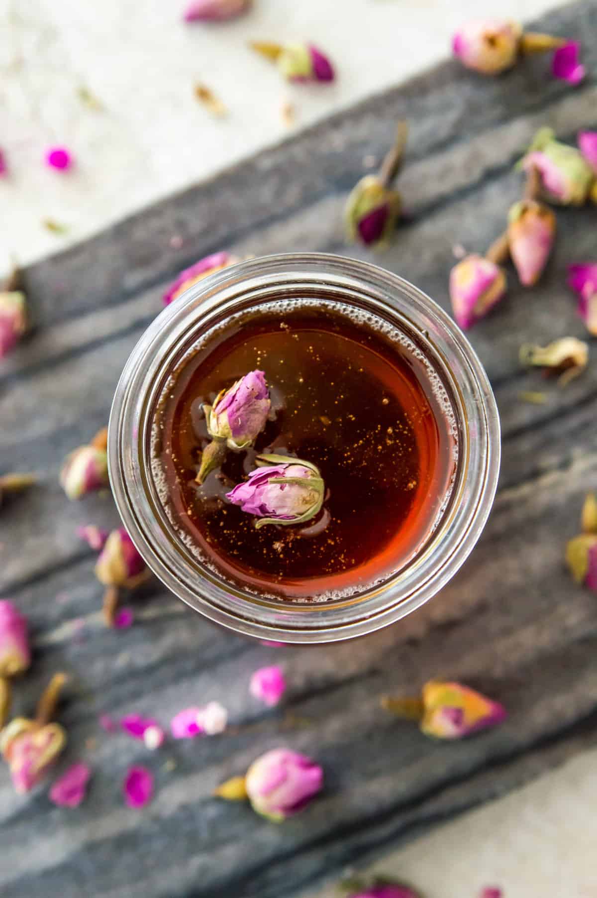 A glass jar filled with a sugar syrup and topped with two dried rose petals.