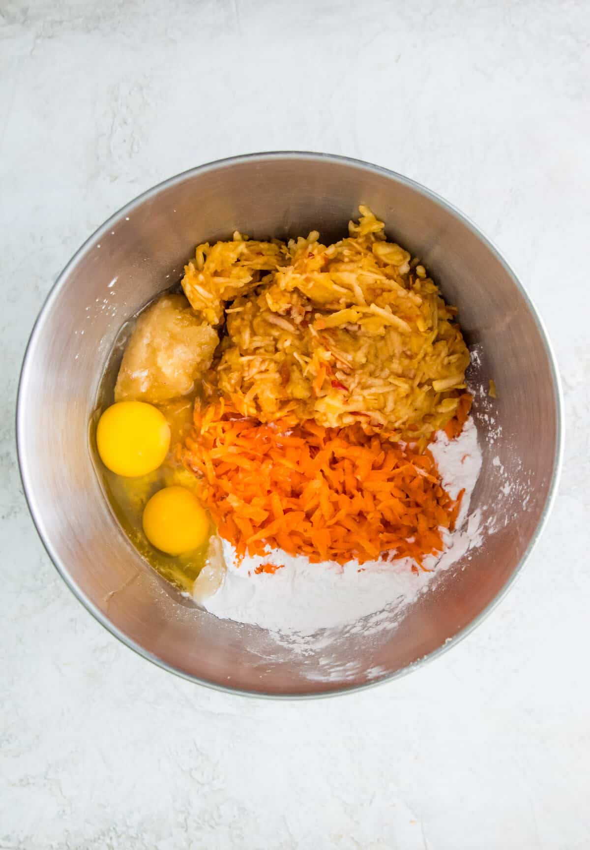 A large stainless steel bowl filled with eggs, shredded carrots, shredded apple, honey and flour.
