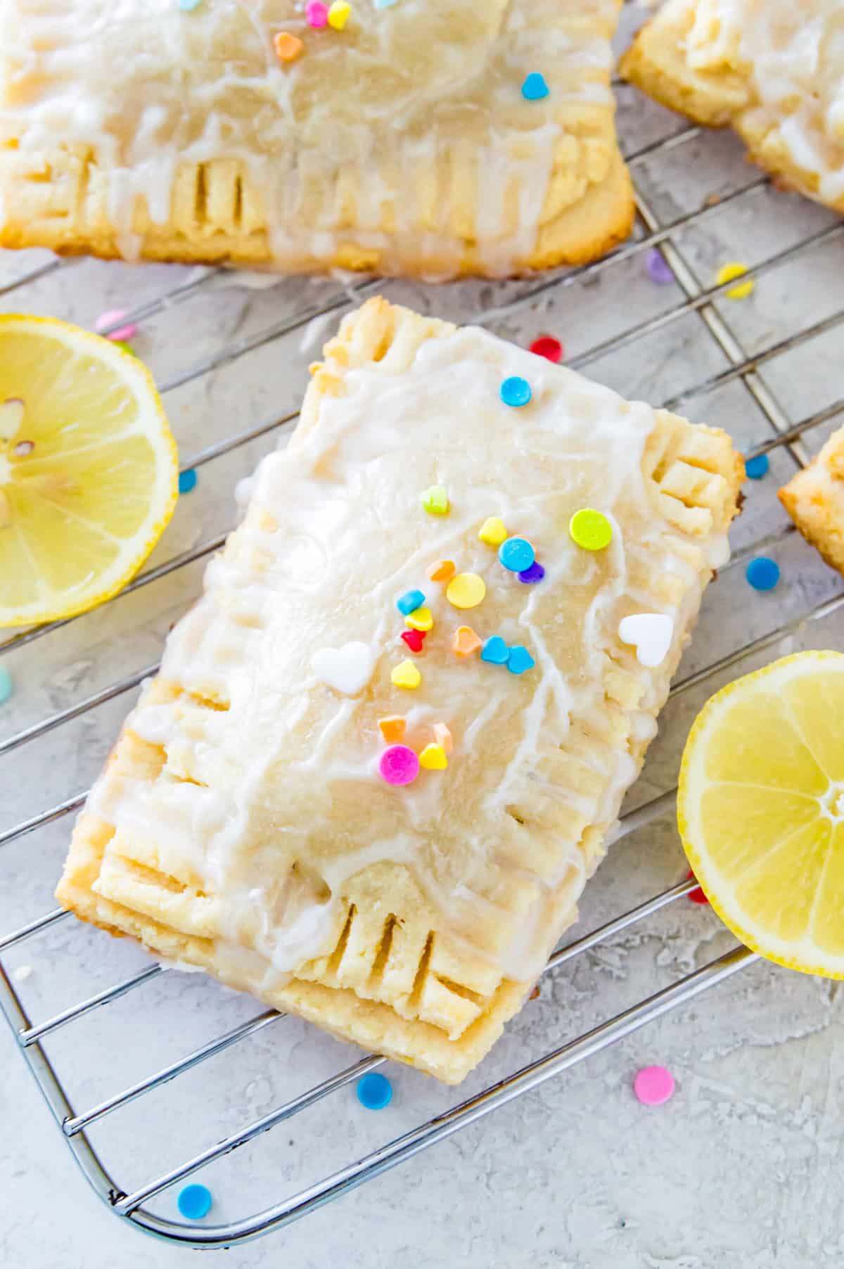 Lemon pop tarts covered in rainbow sprinkles on a wire cooling rack surrounded by lemon slices. 