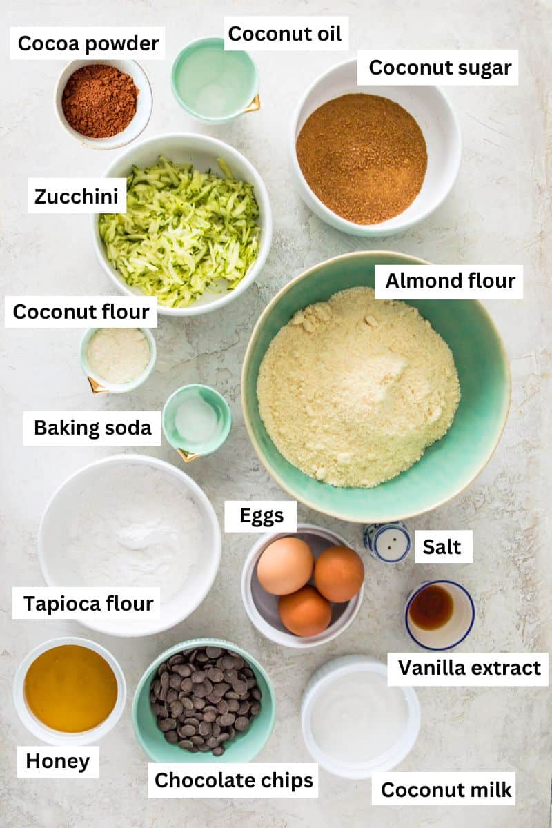 The ingredients needed to make chocolate zucchini bread in separate bowls including flour, cocoa powder, shredded zucchini, eggs and chocolate chips.