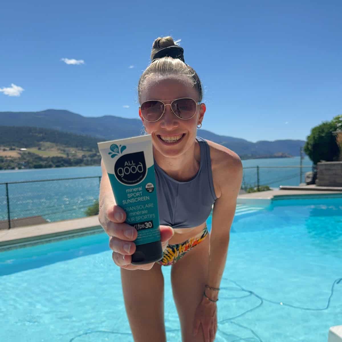 A girl in a bikini in front of a pool holding a bottle of All Good sunscreen. 