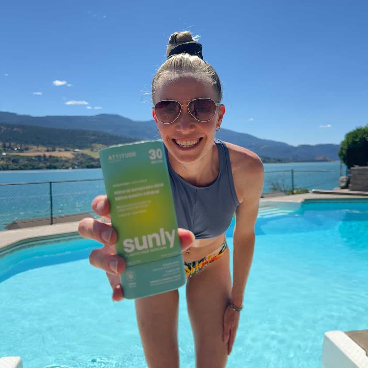 A girl in a bikini in front of a pool holding a tube of Attitude sunscreen.