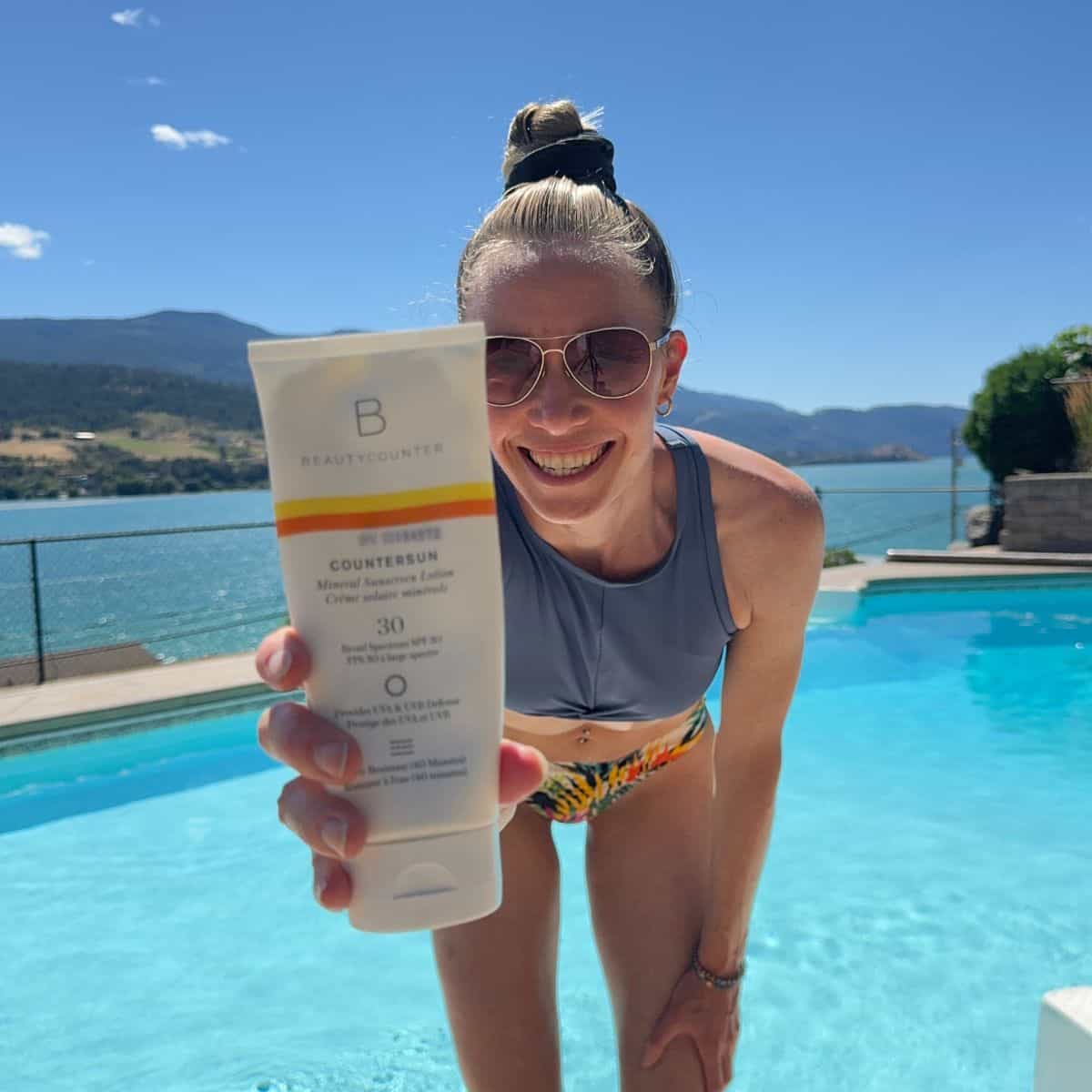 A girl in a bikini in front of a pool holding a bottle of Beautycounter sunscreen.