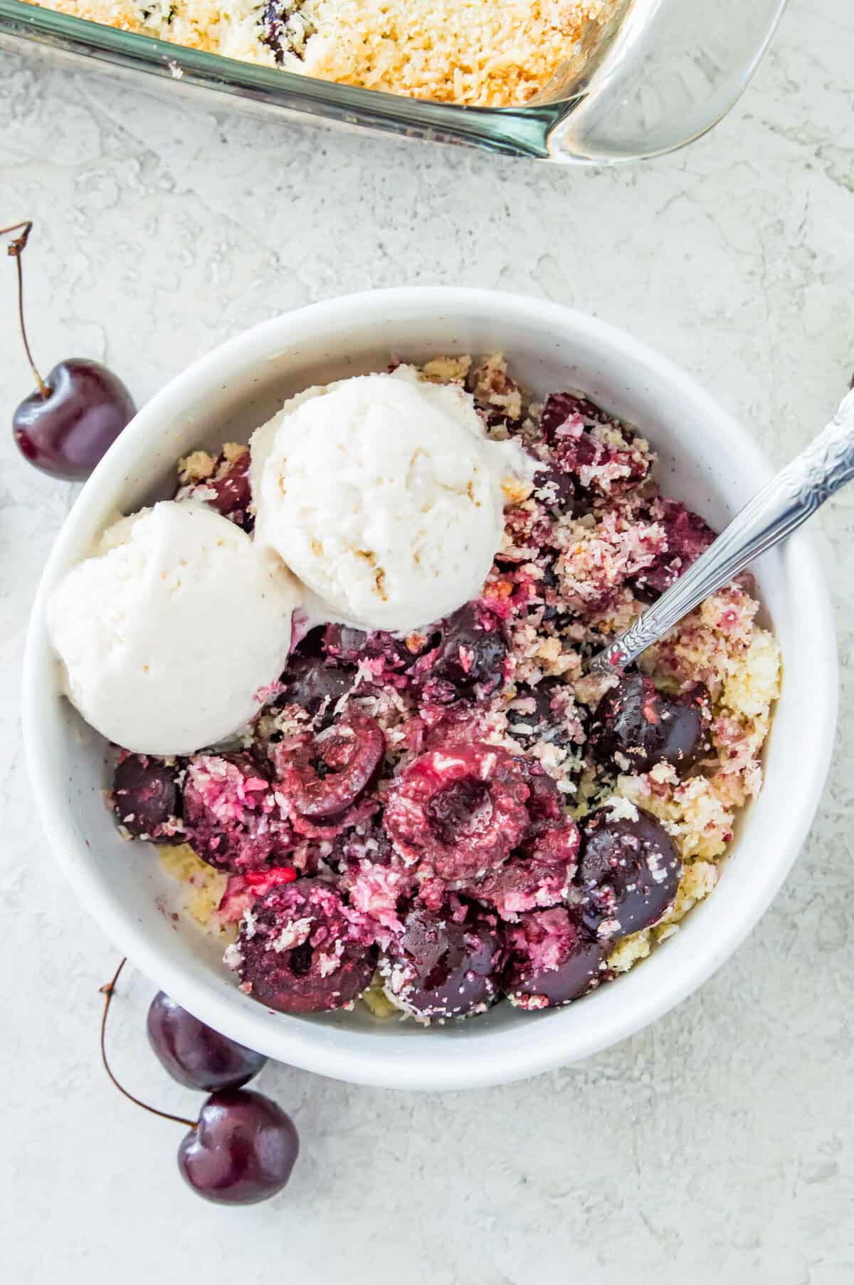 A bowl of cherry crumble with two scoops of vanilla ice cream on it and a spoon in it.